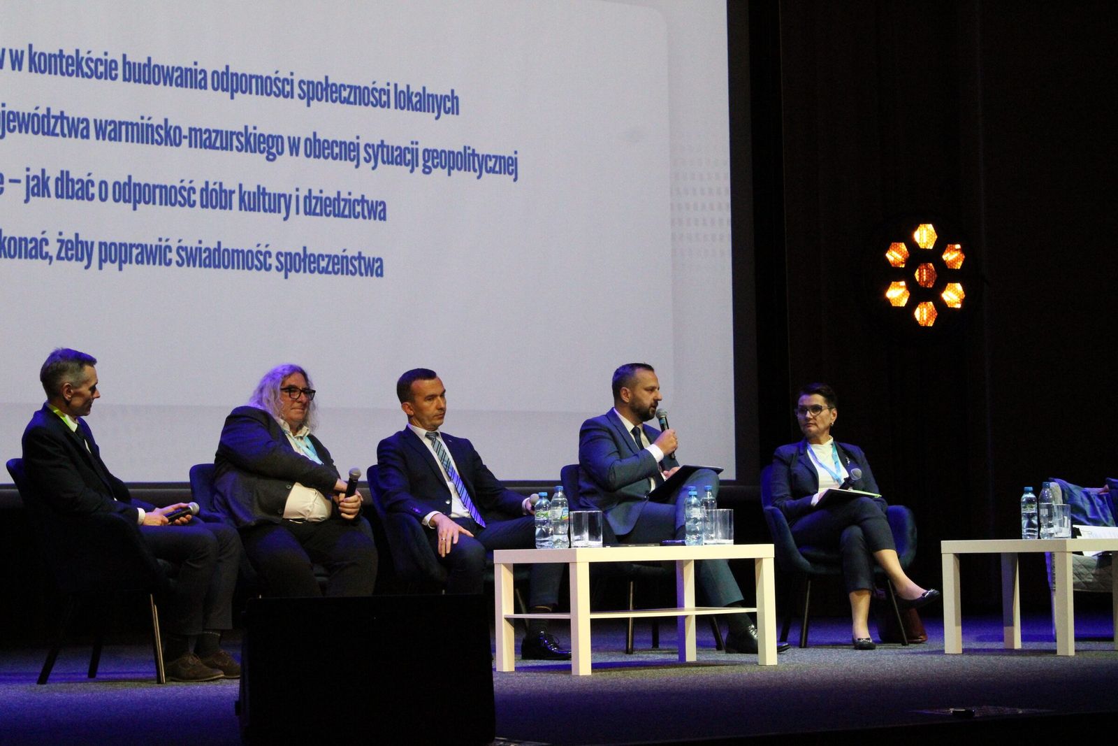 Paneliści na scenie, jeden z nich mówi do mikrofonu.